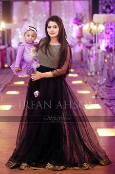 a woman holding a baby wearing a black dress and standing in front of a purple background