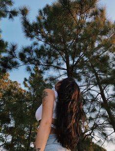 a woman with long hair standing in front of a pine tree looking up at the sky