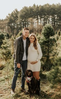 a pregnant couple and their dog pose for a photo in front of some pine trees