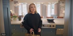 a woman standing in a kitchen holding a brown bag and looking at the camera while wearing a black sweater