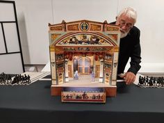 an older man is holding up a model of a building with figurines in front of him