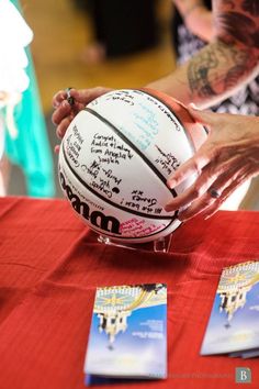 a person holding a basketball on top of a table with other people around it and writing on the ball