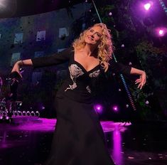 a woman in a long black dress dancing on stage with lights behind her and purple lighting around her