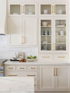 a kitchen with white cabinets and marble counter tops, gold pulls on the glass doors