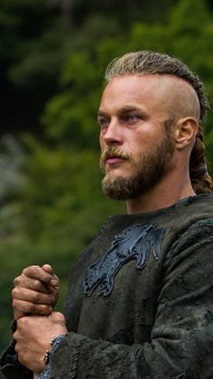 a man with long hair wearing a black shirt and braid standing in front of trees