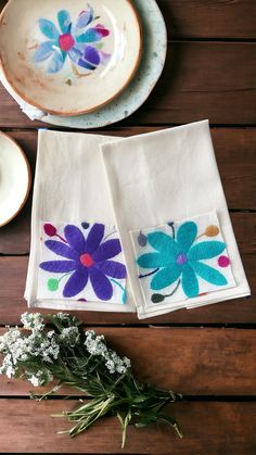 two napkins with flowers painted on them are sitting next to a bowl and plate