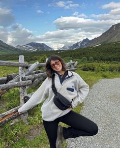 a woman standing on the side of a road next to a wooden fence with mountains in the background