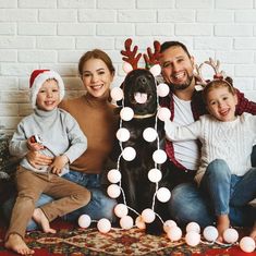 a family sitting on the floor with their dog in front of christmas decorations and lights