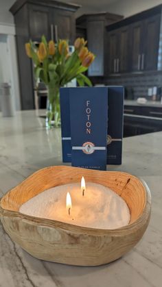 two candles in a wooden bowl on a counter top next to a blue box and flowers