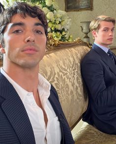 two men in suits sitting on a couch next to each other and one is looking at the camera