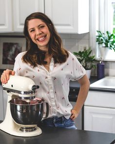 a woman standing next to a kitchen counter with a mixer in front of her and smiling at the camera