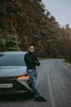 a man standing next to a car on the side of the road with trees in the background