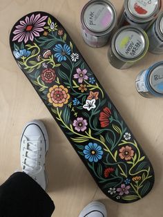 a skateboard with flowers painted on it next to two pairs of shoes and cans