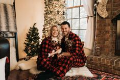 a man and woman sitting on the floor with their dog in front of a christmas tree