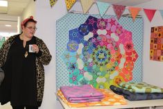 a woman holding a cup in front of some quilts on the wall behind her