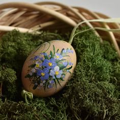 an egg painted with flowers sitting in the grass next to a basket filled with moss