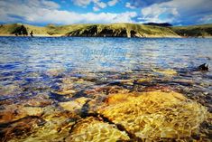 a body of water with rocks in the foreground and mountains in the background, under a cloudy blue sky