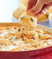 a person dipping some chips into a red bowl filled with cheesy cheese dip