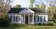 a small white house with green shutters on the front and side windows, surrounded by trees