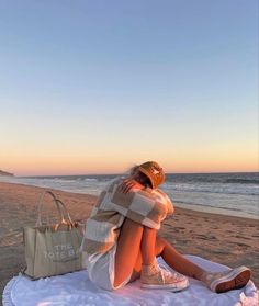 a woman sitting on top of a white blanket next to the ocean with a handbag