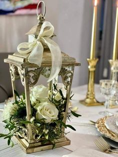 a lantern with flowers and greenery is on a table in front of two candles