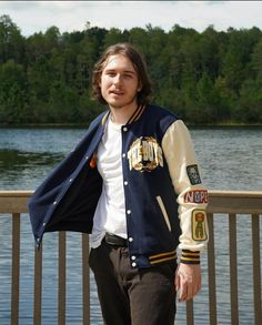 a man with long hair wearing a jacket and pants standing next to a body of water