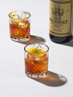 two glasses filled with liquid and ice next to a bottle of old fashioned whiskey on a white surface