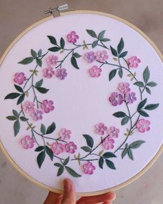 a hand is holding up a white embroidery hoop with pink flowers and green leaves on it