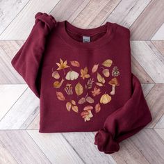 Celebrate autumn in our cozy cottagecore sweatshirt, featuring a heart made of watercolor designs including mushrooms, pinecones, acorns, pumpkins, and fall leaves. This vintage inspired pull-over is great for the nature lover looking to embrace the season's hues and textures. Our crewneck sweatshirts use the highest quality material for ultra-soft and comfortable wear, and they are great for staying snug on chilly days whether you are lounging at home or venturing out. Welcome to Atmos Design C Relaxed Fit Mushroom Print Top For Fall, Relaxed Fit Fall Tops With Mushroom Print, Relaxed Fit Top With Mushroom Print For Fall, Cottagecore Long Sleeve Tops For Fall, Cottagecore Relaxed Fit Top For Fall, Cottagecore Graphic Print Tops For Fall, Winter Cotton Tops With Mushroom Print, Fall Cottagecore Long Sleeve Tops, Cottagecore Cotton Sweater For Fall