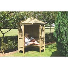 a woman sitting in a wooden gazebo reading a book on the grass next to some trees