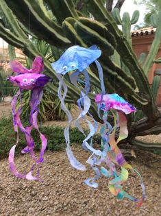 some kind of colorful wind chime hanging from a tree in front of a cactus