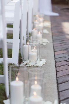 rows of white candles lined up along the side of a brick walkway with petals on the ground