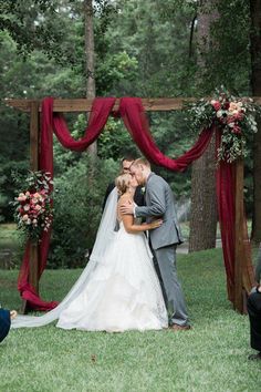 a bride and groom kissing in front of an outdoor ceremony arch with red drapes