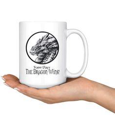 a person holding a white coffee mug with a dragon on it's side and the words, forever day, the dragon wings