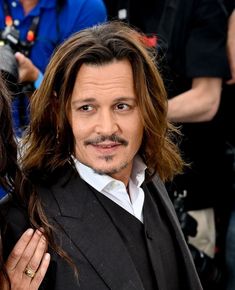 a man with long hair wearing a black suit and tie next to a woman in a white shirt