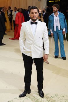 a man in a white tuxedo and bow tie standing on a carpet with other people