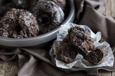 two chocolate donuts sitting on wax paper next to a bowl of doughnuts