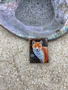 a small square tile with a red fox on it's side in front of a bowl full of white rice