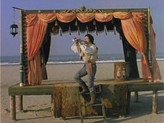 a man standing on top of a wooden bench next to an open stage covered in orange drapes