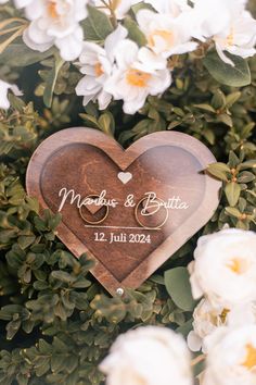 a wooden heart with two wedding rings on it surrounded by white flowers and greenery