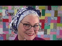 an older woman wearing glasses and a head scarf smiles in front of a quilted wall