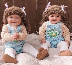 two babies sitting on top of a bed wearing overalls and knitted hats with bows