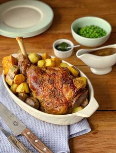 a roasting pan filled with meat, potatoes and peas next to a knife on a wooden table