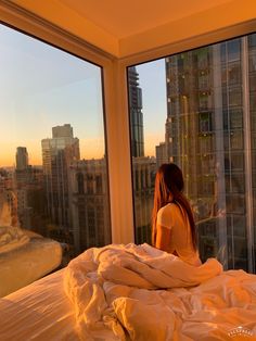 a woman sitting on top of a bed in front of a window looking out at the city