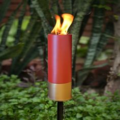 a red candle with yellow flames in the middle of some bushes and trees behind it