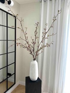 a white vase with flowers in it sitting on a black stand next to a mirror