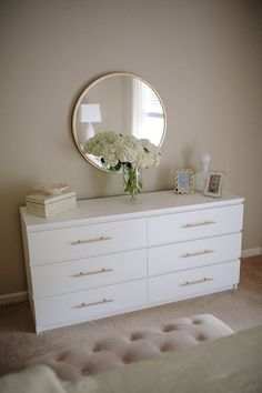 a white dresser with flowers on top and a round mirror above it in a bedroom