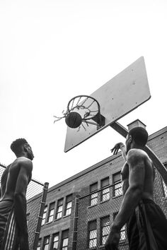 two young men playing basketball in front of a large brick building with an overhead basketball hoop