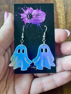 a pair of blue ghost earrings with purple splats on them, sitting in front of a card