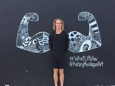 a woman standing in front of a black wall with white and gray drawings on it